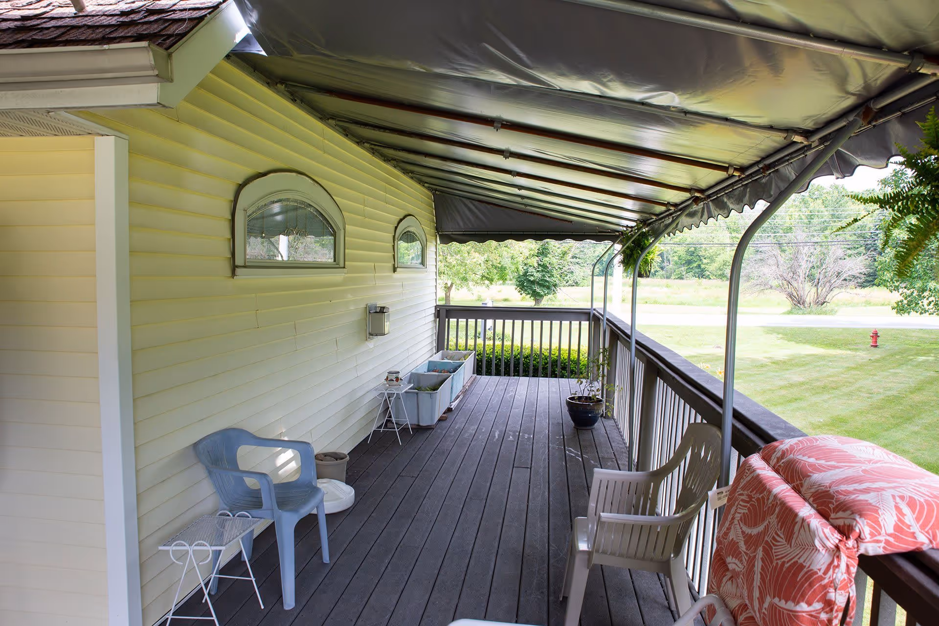 Covered wooden porch with plastic chairs, potted plants, and railing overlooking a grassy lawn.