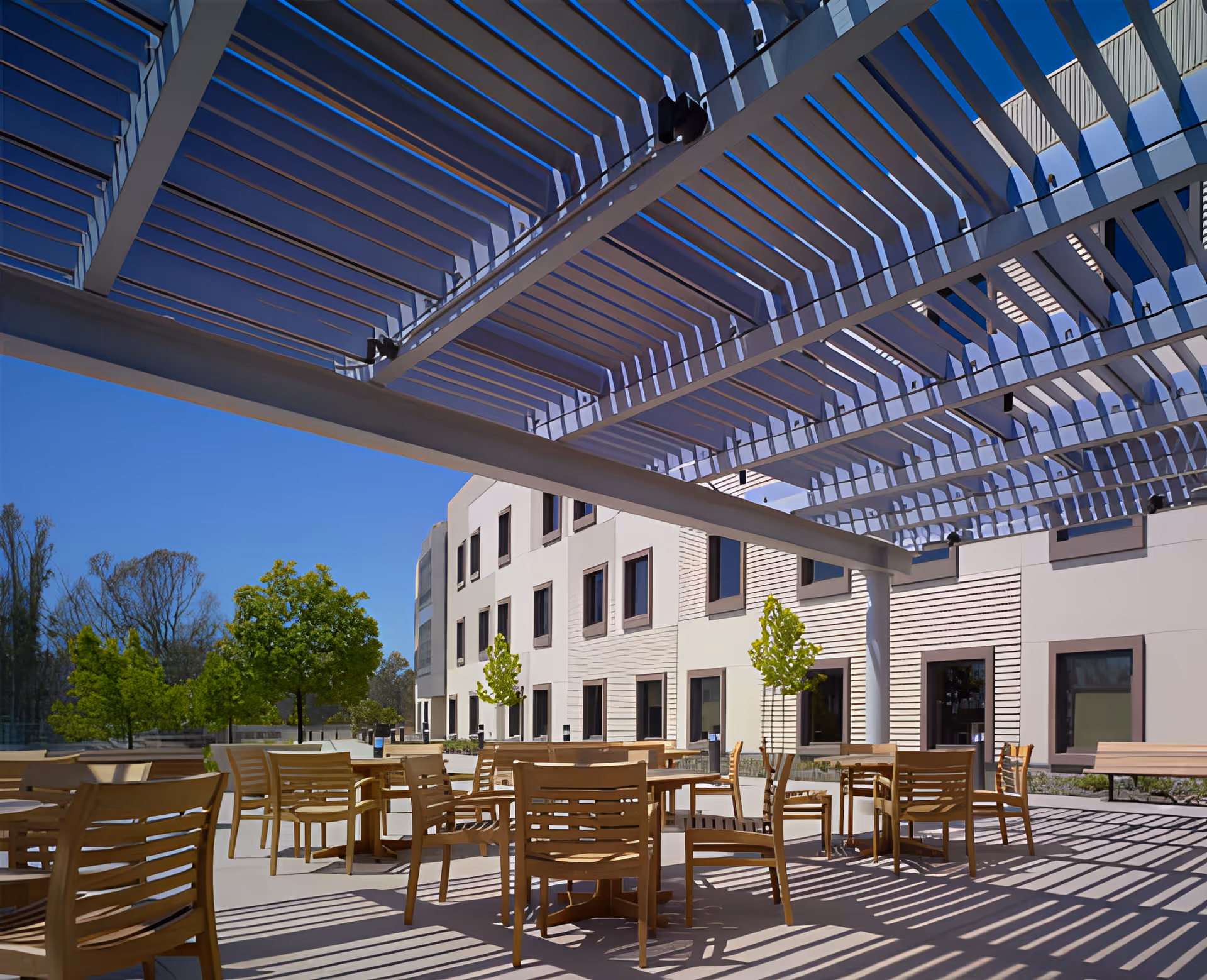 Shaded outdoor patio with wooden tables and chairs under a large slatted pergola beside a multi-story building.