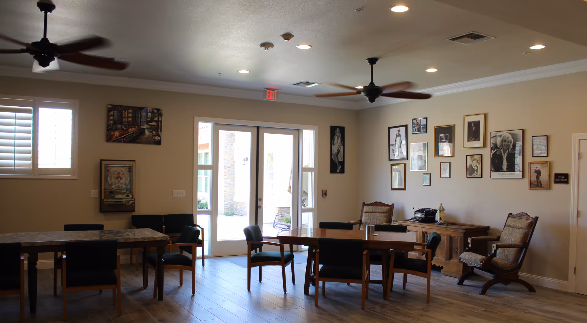 A well-lit common room with wooden floors, two ceiling fans, and multiple tables and chairs arranged for seating. The walls are decorated with framed black and white photographs, including images of Marilyn Monroe. There is a door leading outside with glass panels on either side, and a sign on the wall indicating a staff restroom.