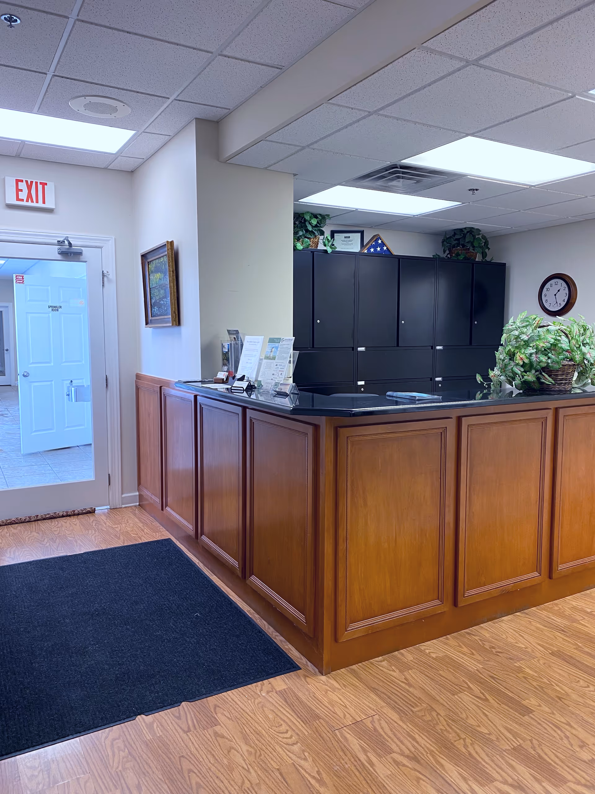 Interior reception desk with dark cabinets, plants, clock, and an exit door.