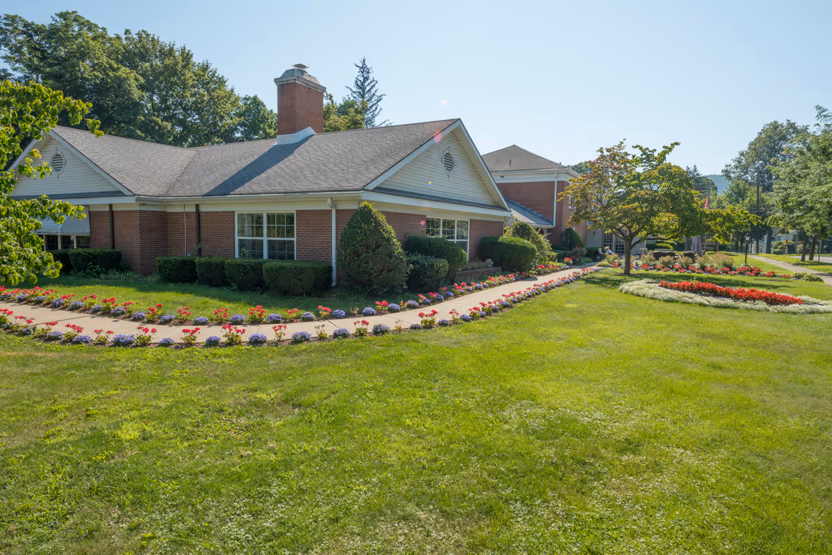 Single-story brick senior living facility with manicured lawn and flower beds along a curved walkway.