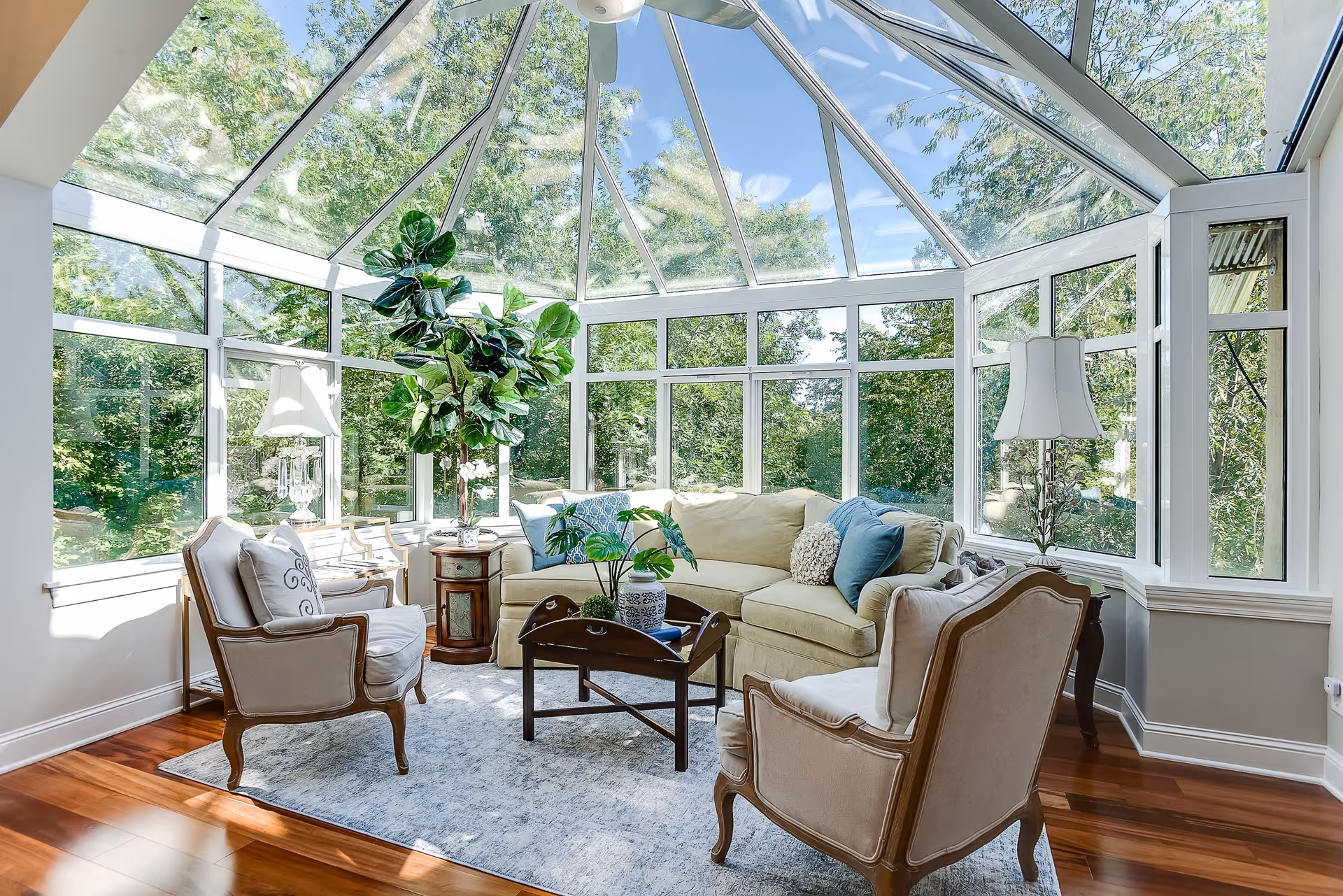 A bright sunroom with large glass windows and a glass ceiling, surrounded by green trees outside. Inside, there is a beige sofa with blue and beige cushions, two beige armchairs with wooden frames, a wooden coffee table with plants on it, a small wooden side table with a lamp, and a light-colored area rug on a polished wooden floor.
