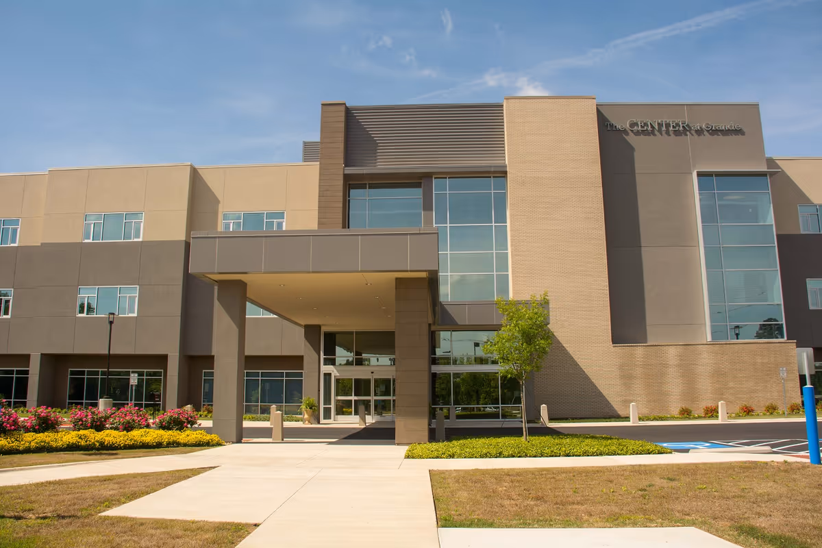 Exterior front view of The Center At Grande building with a covered entrance, large windows, and landscaped surroundings including flowers and a small tree under a clear blue sky.