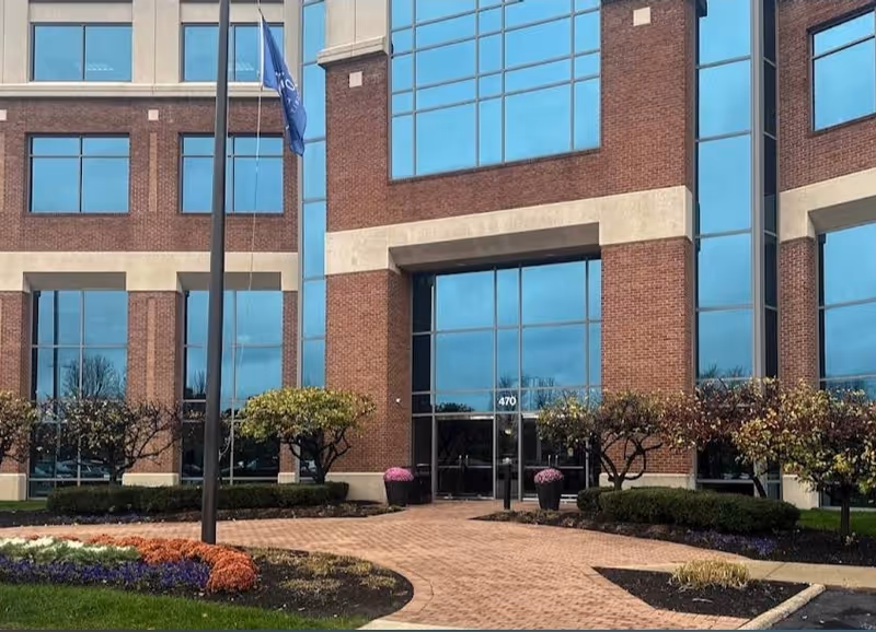 Brick building exterior with large glass windows, flagpole, and landscaped entrance walkway.