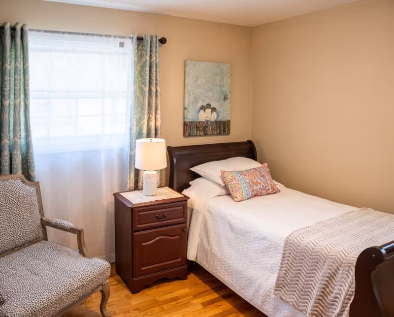 A cozy bedroom with a single bed, nightstand, lamp, chair, and window.