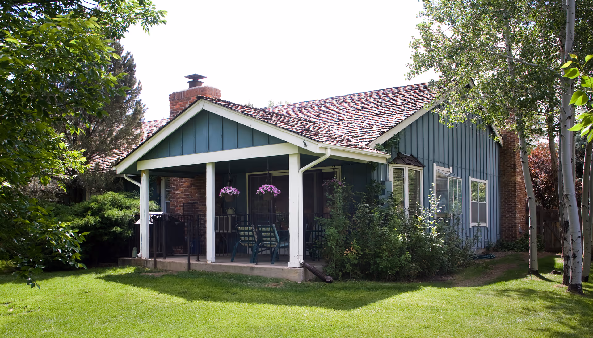 A single-story blue house with a covered porch featuring white pillars, hanging flower pots, and outdoor chairs. The house has a shingled roof, a brick chimney, and is surrounded by green grass, bushes, and trees.
