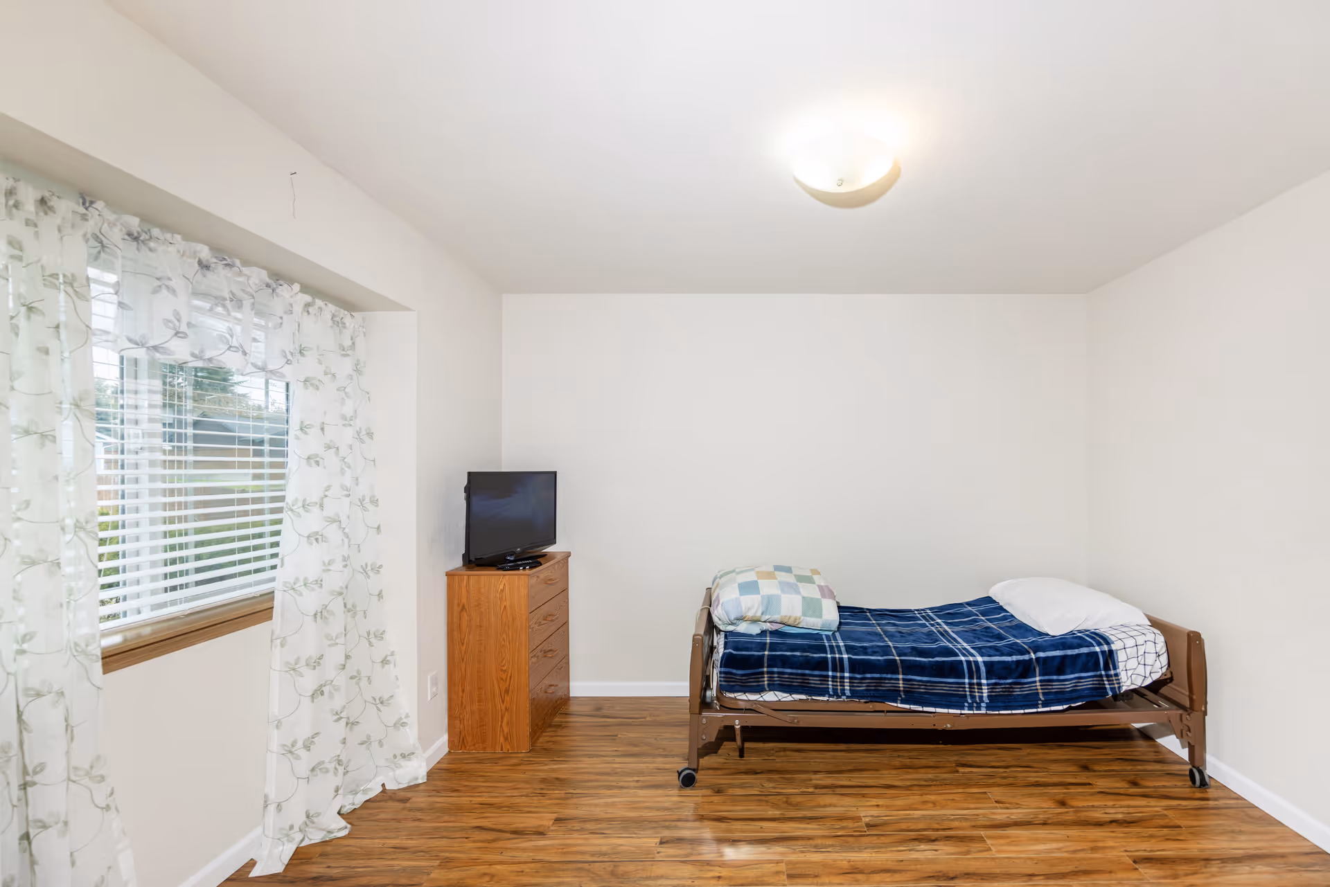 A simple bedroom in a senior living facility with a single bed, dresser, and TV.
