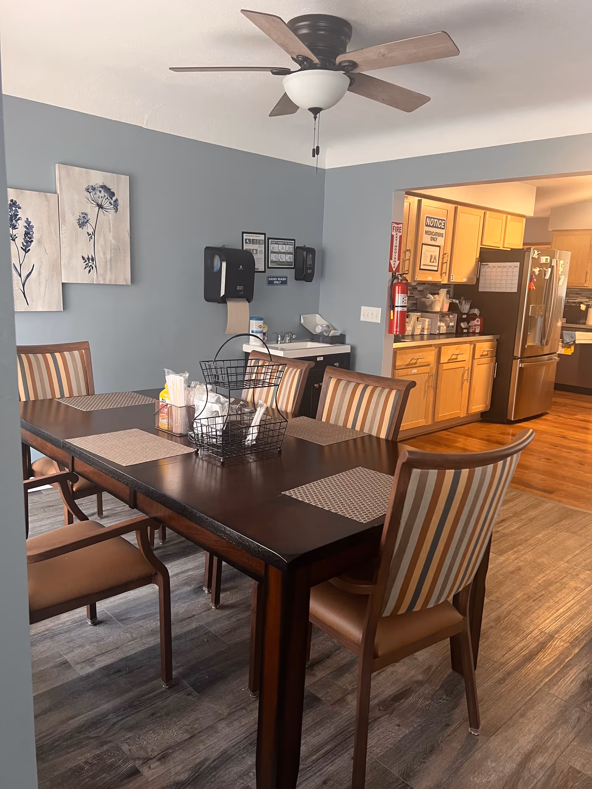 Dining room with a dark table, striped chairs, wall decor, and a kitchenette in the background.