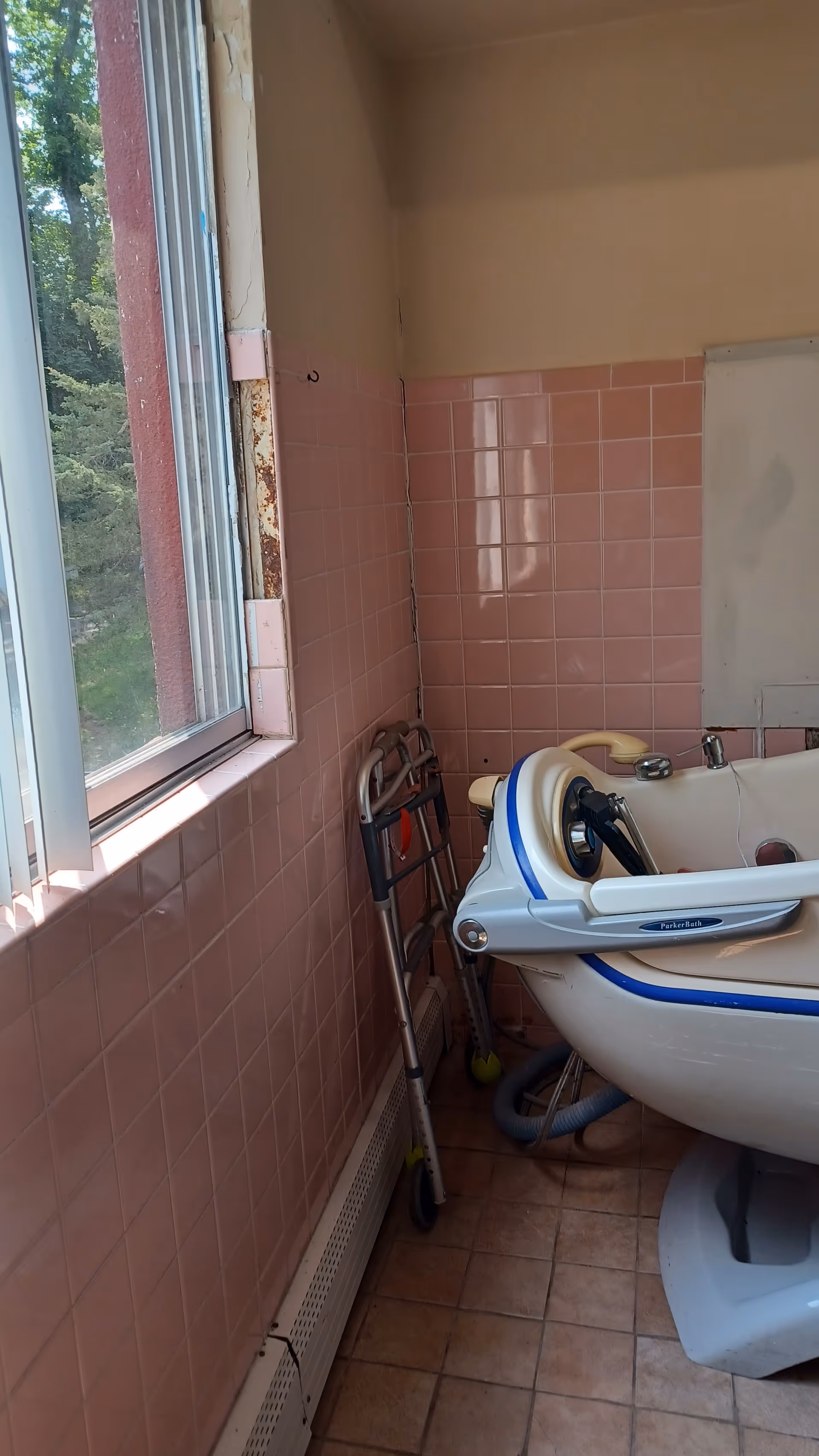 Pink-tiled bathroom with a bathtub lift and a walker beside a sunlit window.