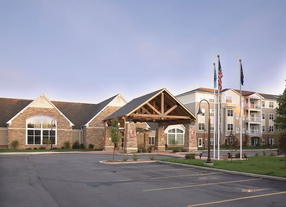 Exterior view of a senior living facility with a large covered entrance supported by stone pillars and wooden beams. The building has multiple floors with balconies and large windows. Three flagpoles with flags are positioned near the entrance, and there is a parking lot in the foreground.