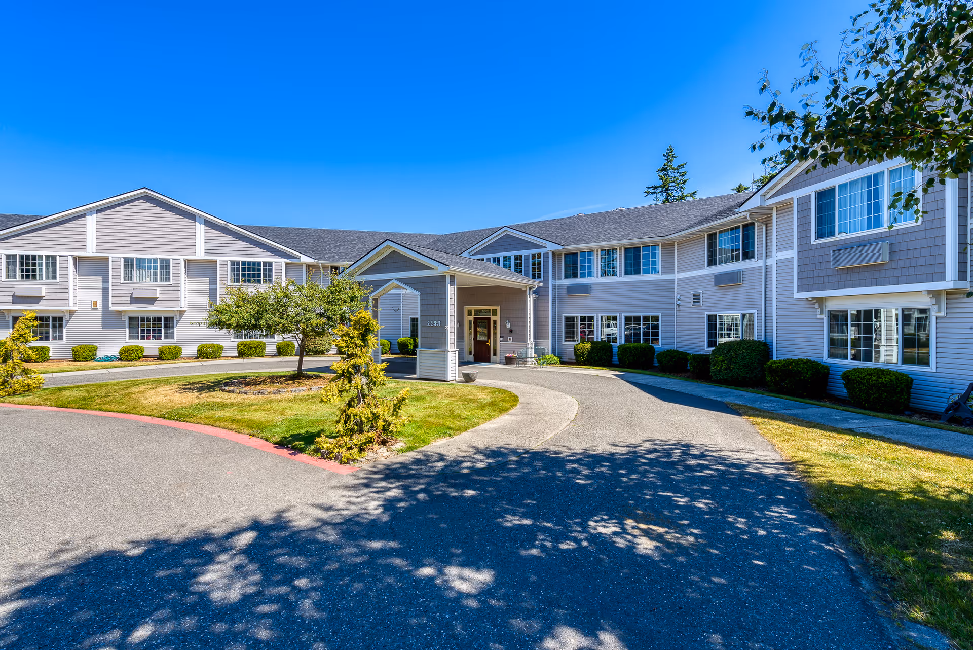 Front entrance and driveway of a light-gray two-story assisted living building with landscaped lawn under a clear blue sky.