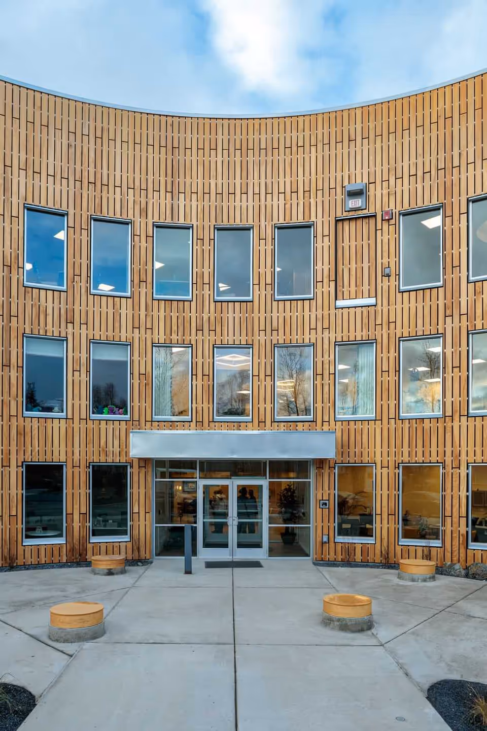 Front entrance of a curved, wood-clad building with multiple windows and a glass double door.