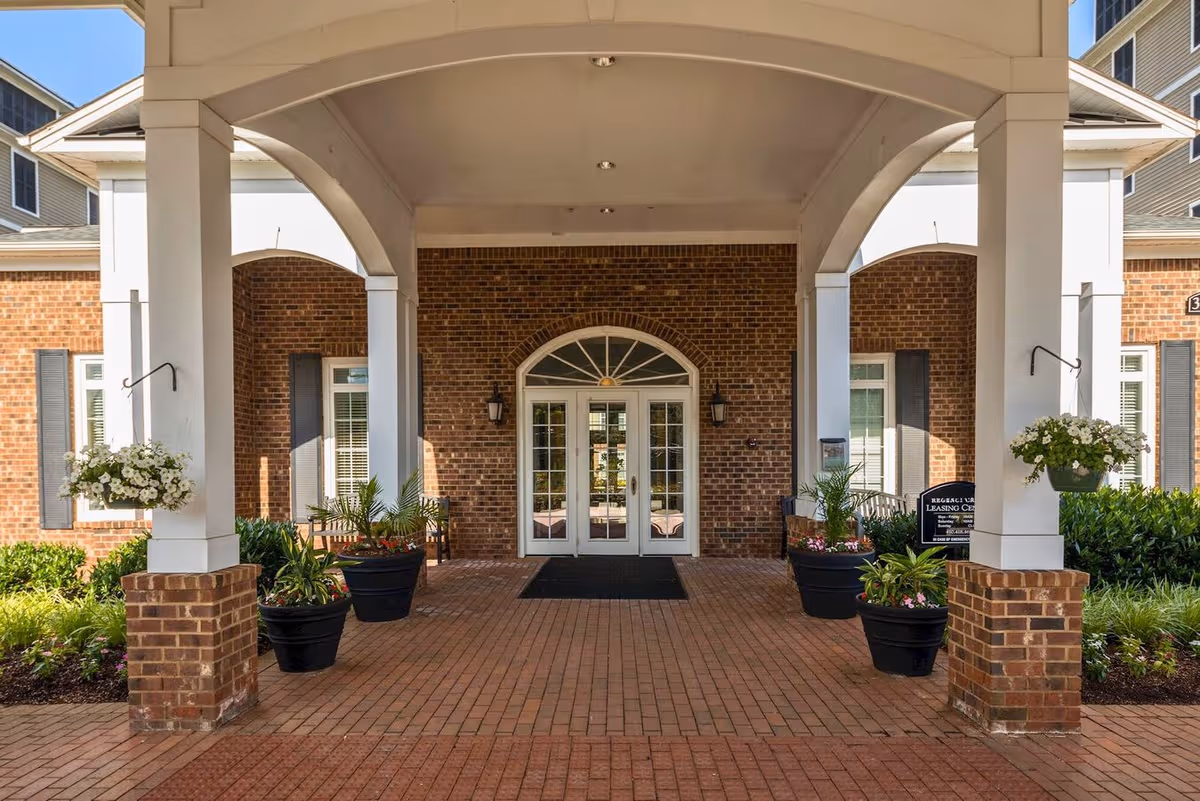 Brick senior living building entrance with arches, columns, and potted plants.