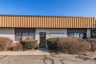 Single-story building with a metal awning, entry door, and trimmed bushes.