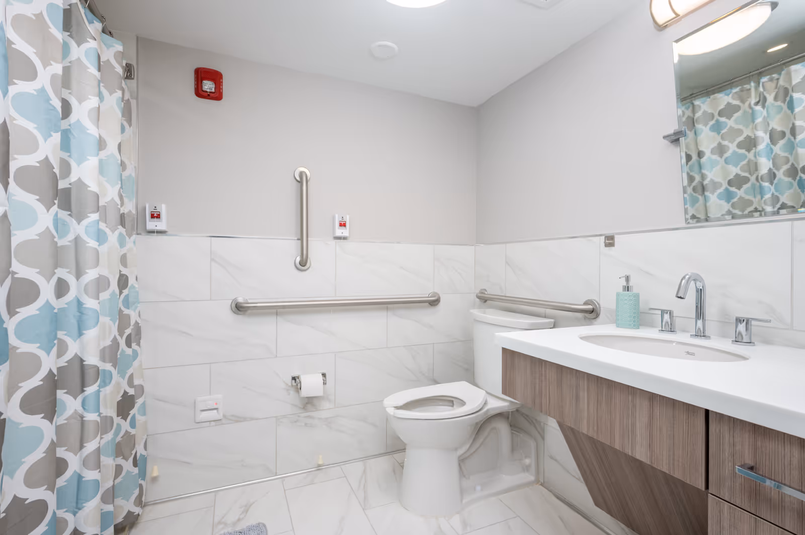 Accessible bathroom with grab bars, toilet, sink, and patterned shower curtain.
