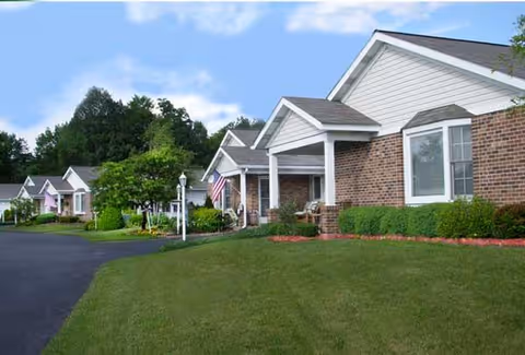 Row of single-story brick cottages with small porches, landscaped lawns, and an asphalt driveway.