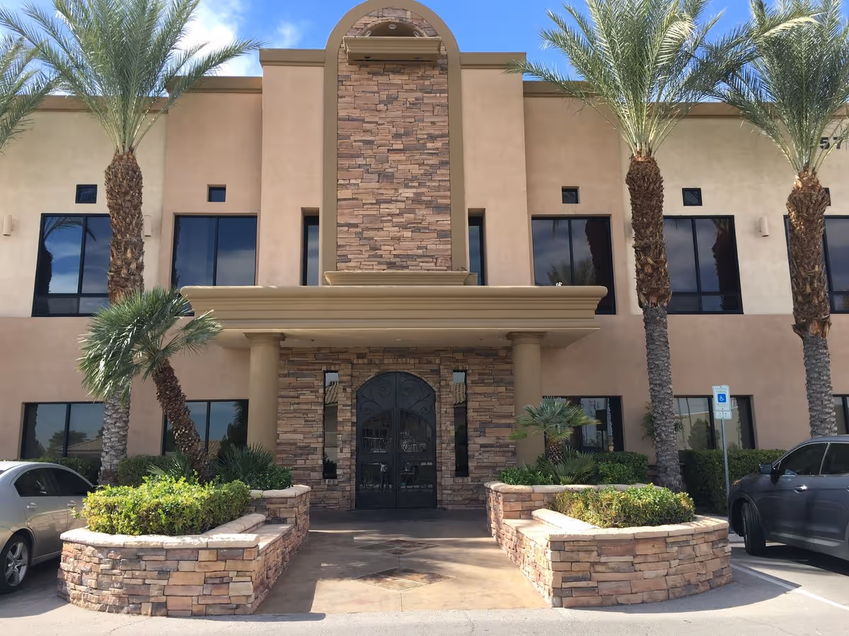 Exterior view of a beige senior living facility with stone facade, arches, and palm trees.