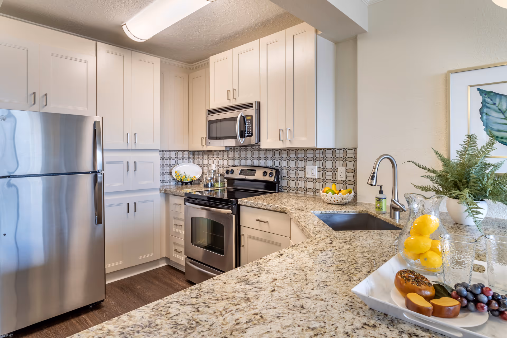 Modern kitchen with white cabinets, stainless steel refrigerator, oven, and microwave. Granite countertops with a sink and faucet, decorative backsplash, and a tray with lemons, glasses, and fruit on the counter. A potted plant and framed artwork are on the wall.