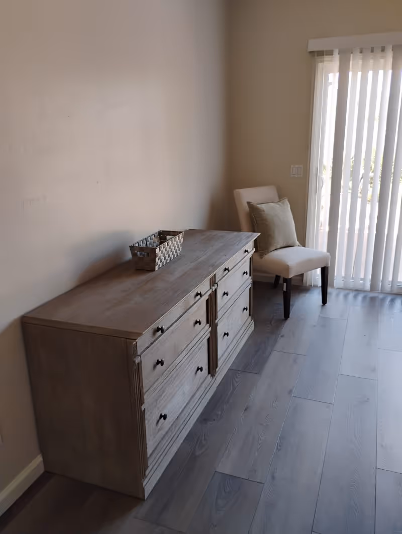 A wooden dresser with multiple drawers and black knobs is placed against a beige wall. On top of the dresser is a small woven basket. To the right of the dresser is a beige upholstered chair with a cushion, positioned near a sliding glass door with vertical blinds, allowing natural light to enter the room. The floor is covered with light gray wood-like planks.