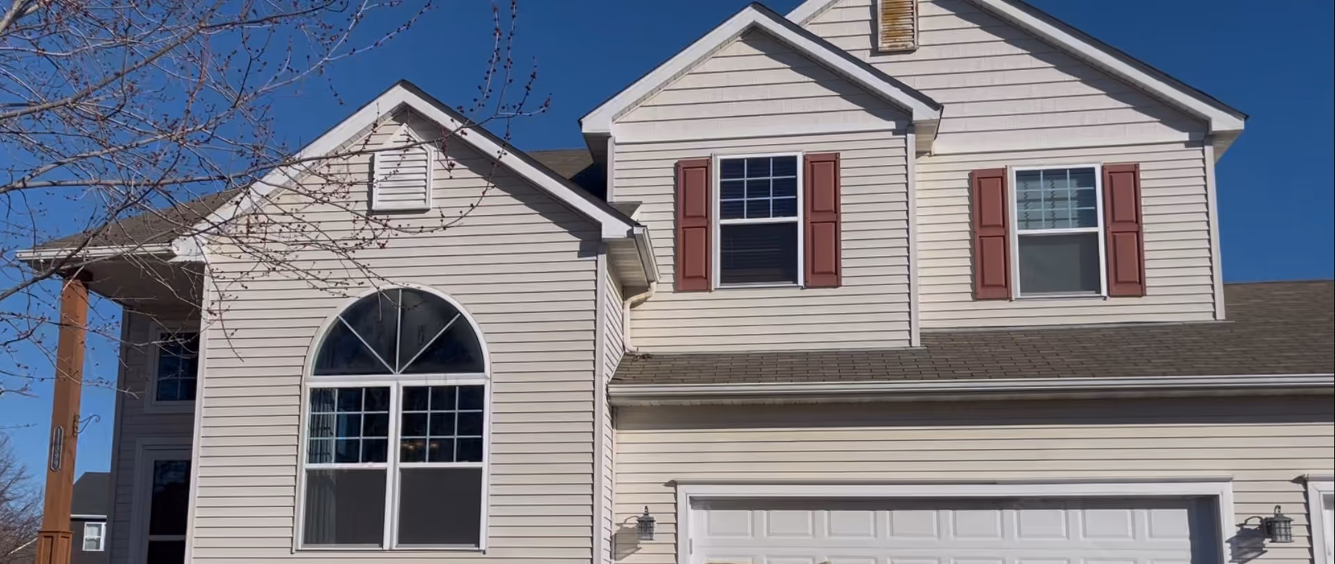 Two-story beige house with arched front window, red shutters, garage, under blue sky.