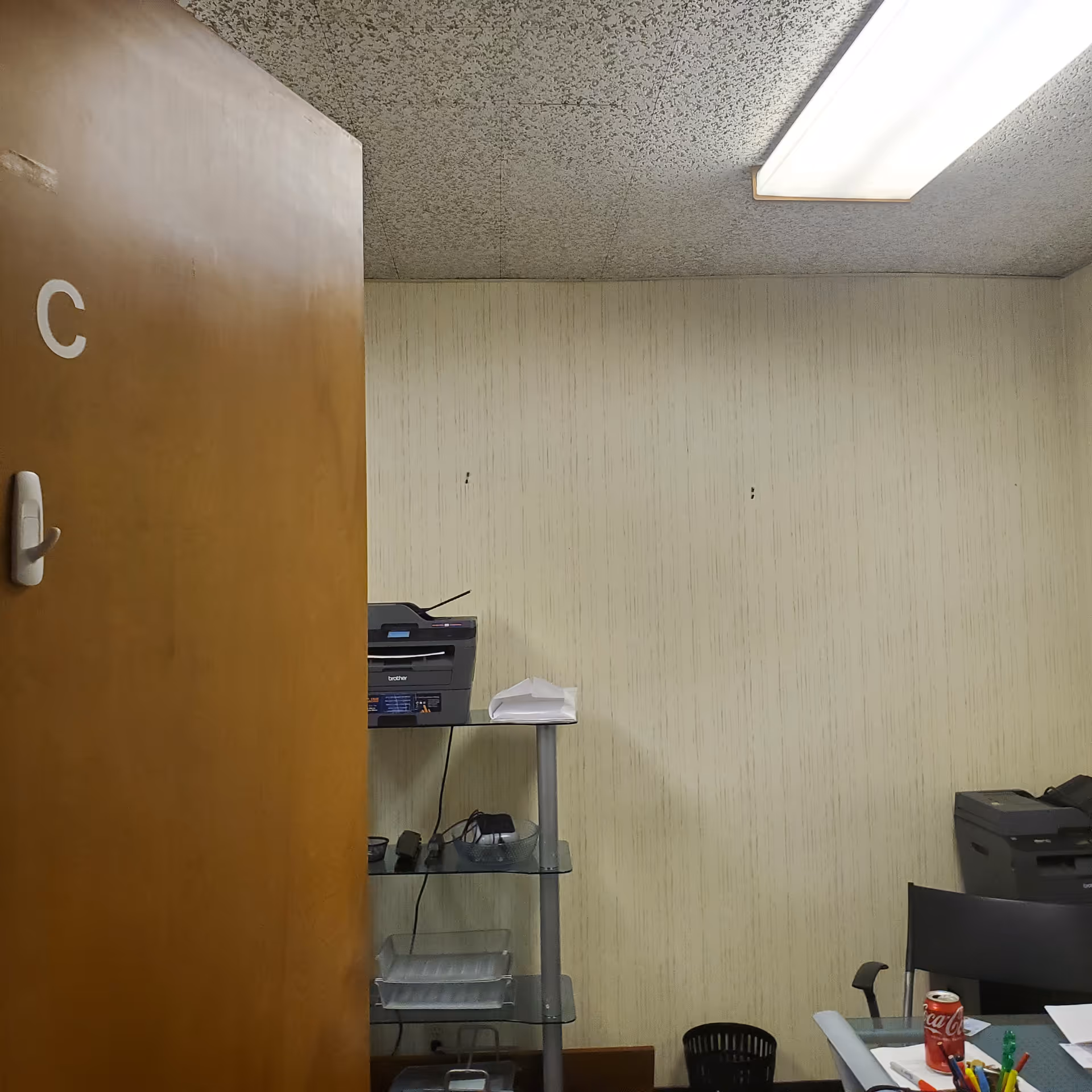 Office corner inside facility with door marked C, printer, shelves, paperwork, and a soda can.
