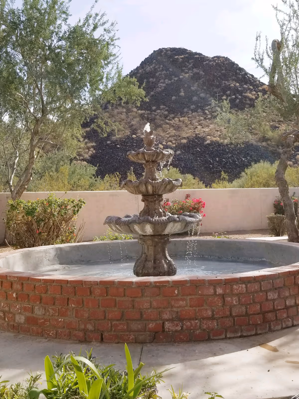 A three-tiered stone water fountain surrounded by a circular red brick wall in an outdoor garden area with trees, bushes, and flowers. In the background, there is a rocky hill under a clear sky.