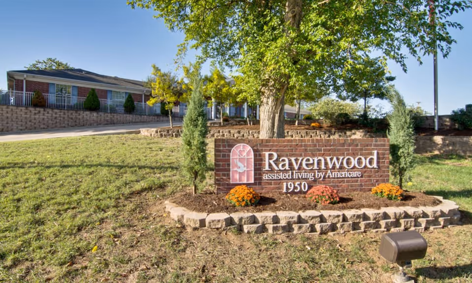 Outdoor view of Ravenwood Senior Living facility with a brick sign displaying the name 'Ravenwood assisted living by Americare 1950' surrounded by small trees and flowers, with the building and a clear blue sky in the background.