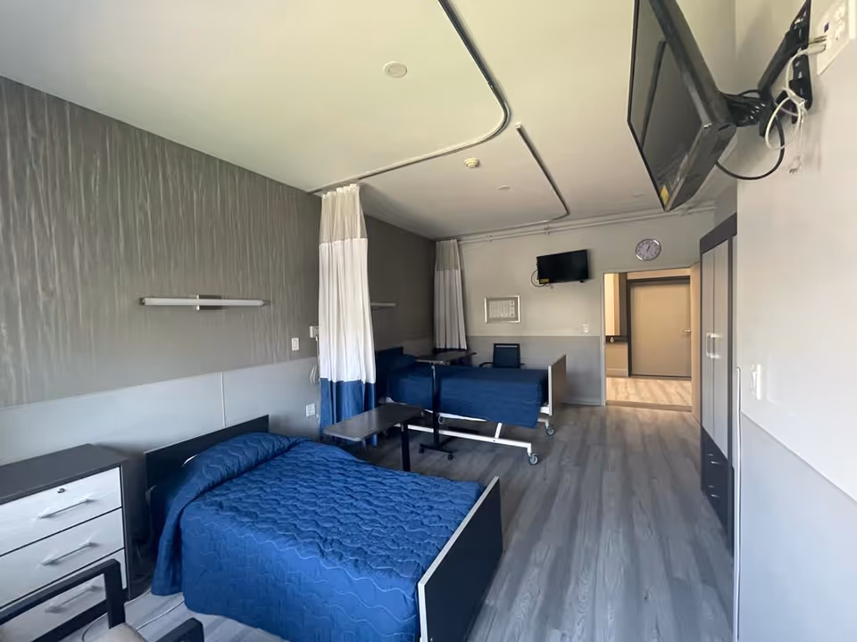 A nursing center patient room with two blue beds, curtains, and a wall-mounted TV.