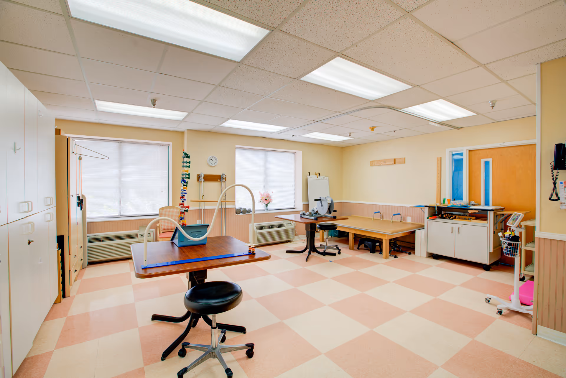 Bright, empty senior care room with desks, chairs, cabinets, medical equipment, and treatment tables.