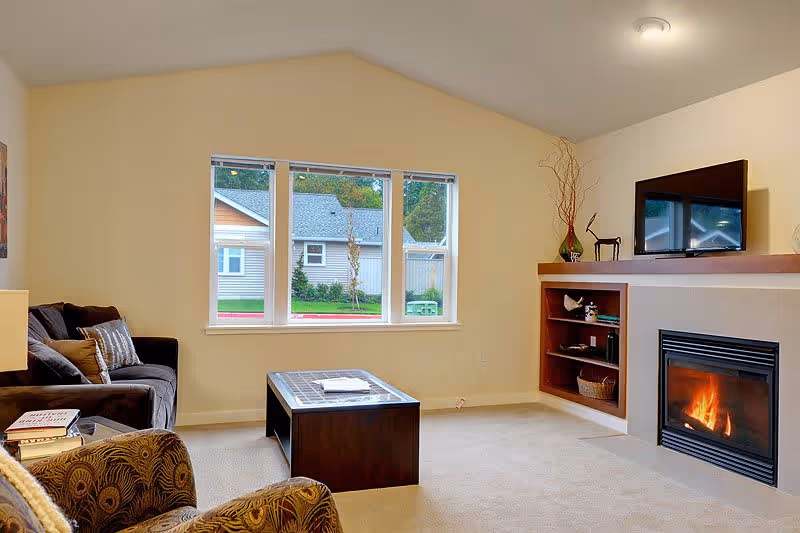 Living room with sofa and armchairs, a coffee table, TV above a built-in fireplace, and a window overlooking neighboring houses.