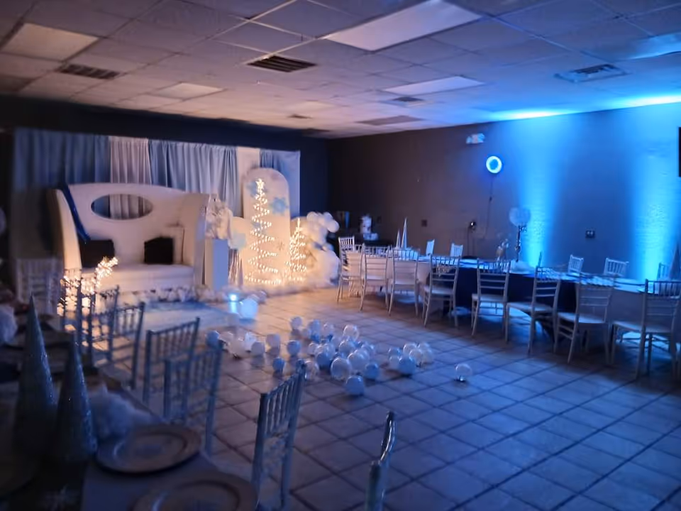 Blue-lit indoor dining area with round tables, chairs, and a white stage backdrop.
