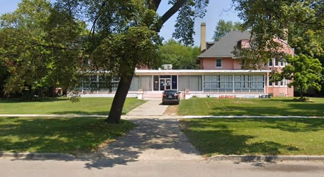 Front view of a pink senior living facility with a long enclosed porch, lawn, sidewalk, trees, and a parked car.
