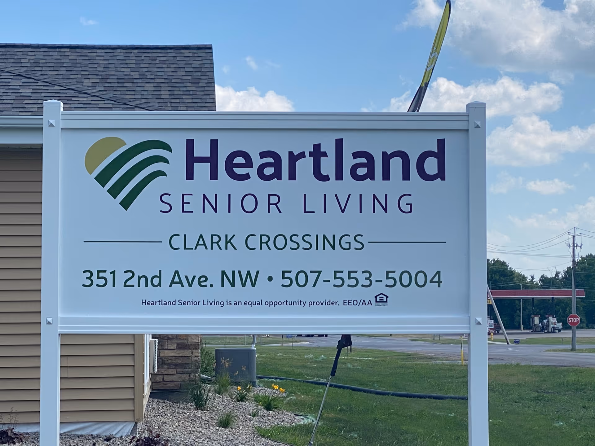 Outdoor sign for Heartland Senior Living at Clark Crossings, displaying the address 351 2nd Ave. NW and phone number 507-553-5004, with a building and grassy area in the background under a partly cloudy sky.