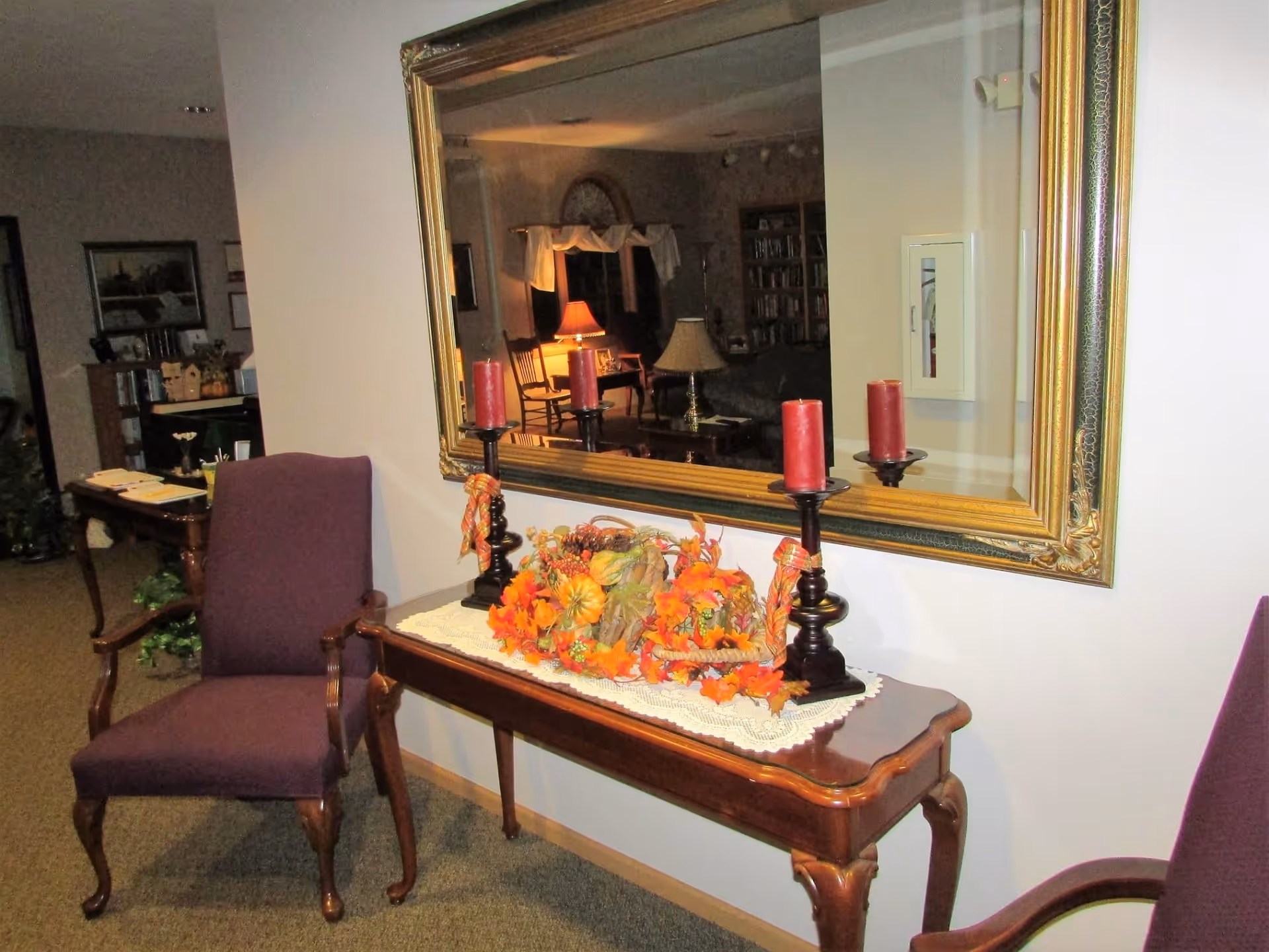 A hallway area in a retirement community featuring a wooden console table with a lace runner, decorated with autumn-themed foliage and three red candles on black candle holders. Two purple upholstered chairs with wooden armrests flank the table. A large ornate gold-framed mirror hangs on the wall above the table, reflecting a cozy living room with lamps, chairs, and bookshelves.