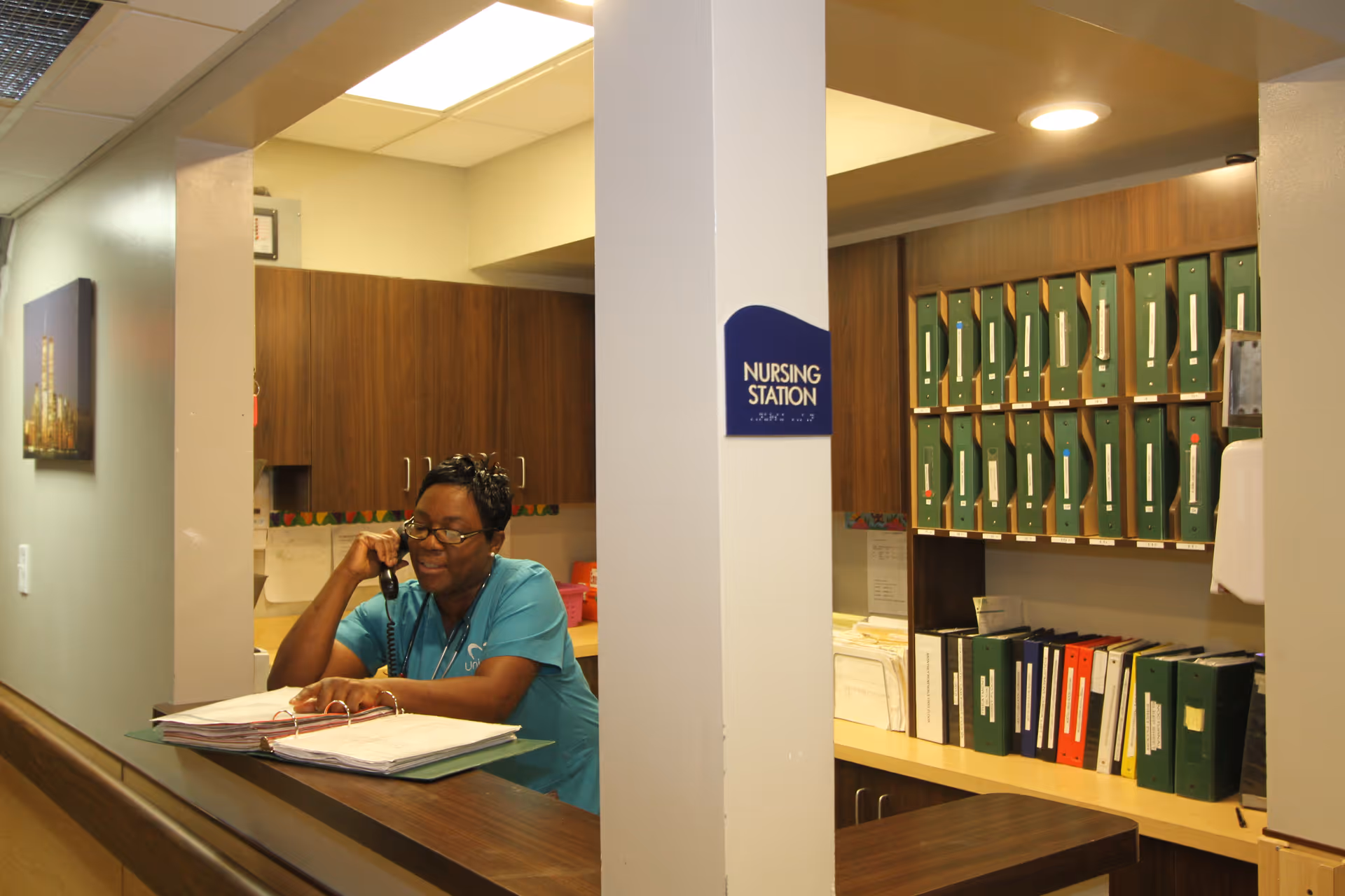 A nurse in teal scrubs is sitting at a nursing station desk, talking on the phone and looking at an open binder. The nursing station has wooden cabinets and shelves filled with green and other colored binders and folders.