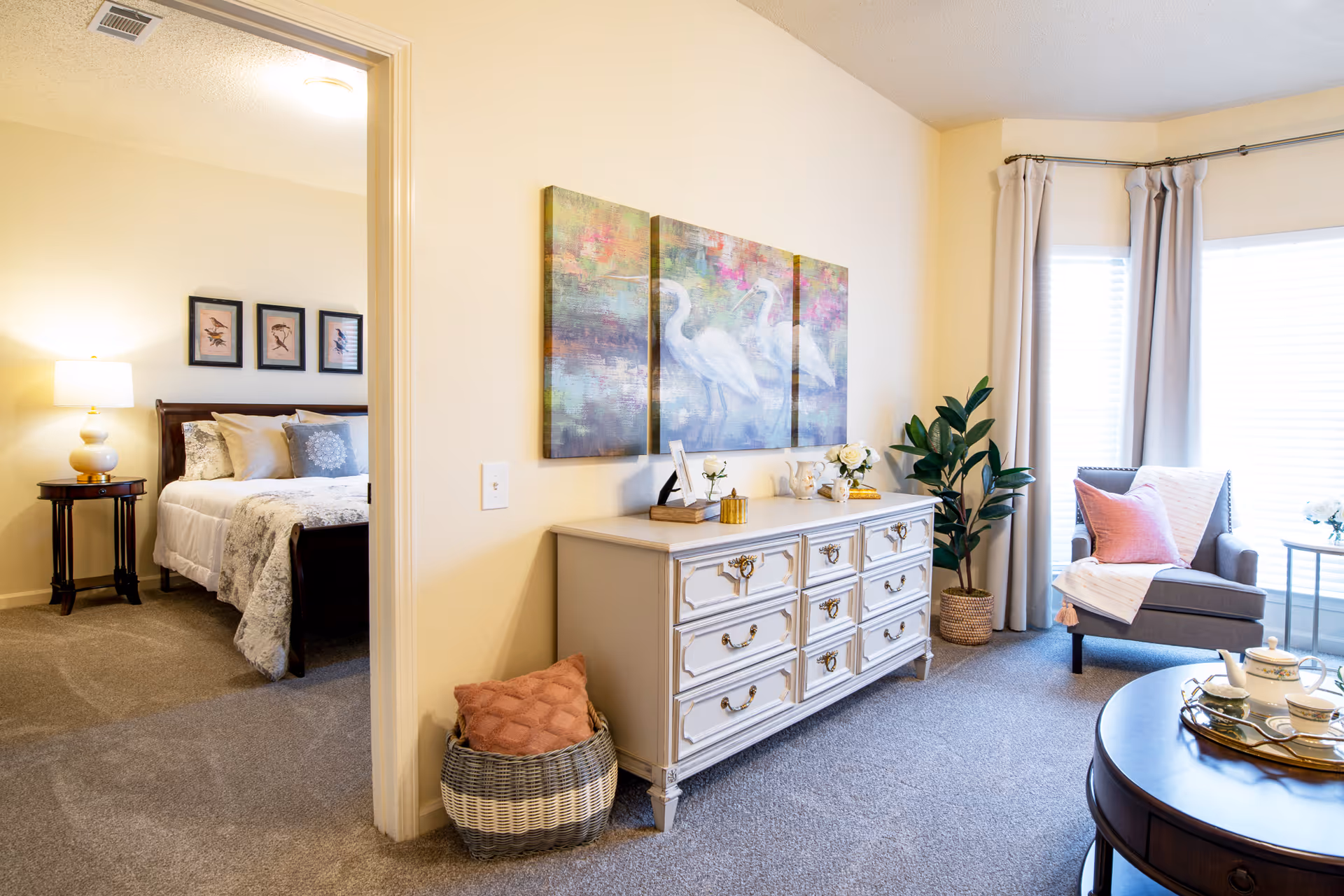 A cozy living area with a gray armchair draped with a pink pillow and a white throw blanket, a round wooden coffee table with a tea set, and a white dresser adorned with decorative items and a three-panel painting of white birds on the wall above. To the left, an open doorway reveals a bedroom with a bed, pillows, a bedside table, and a lamp. The room is carpeted and well-lit with natural light coming through windows with white blinds and beige curtains.