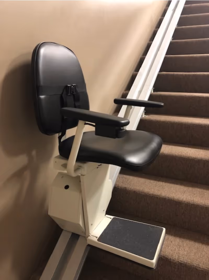 Indoor stair-lift chair mounted beside carpeted stairs in a hallway at a senior living facility.