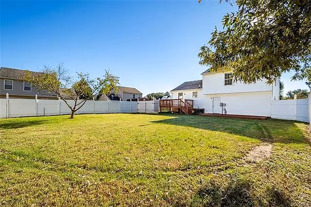 Fenced backyard with a green lawn, a small tree, and the rear of a white two-story house with a wooden deck under a clear blue sky.