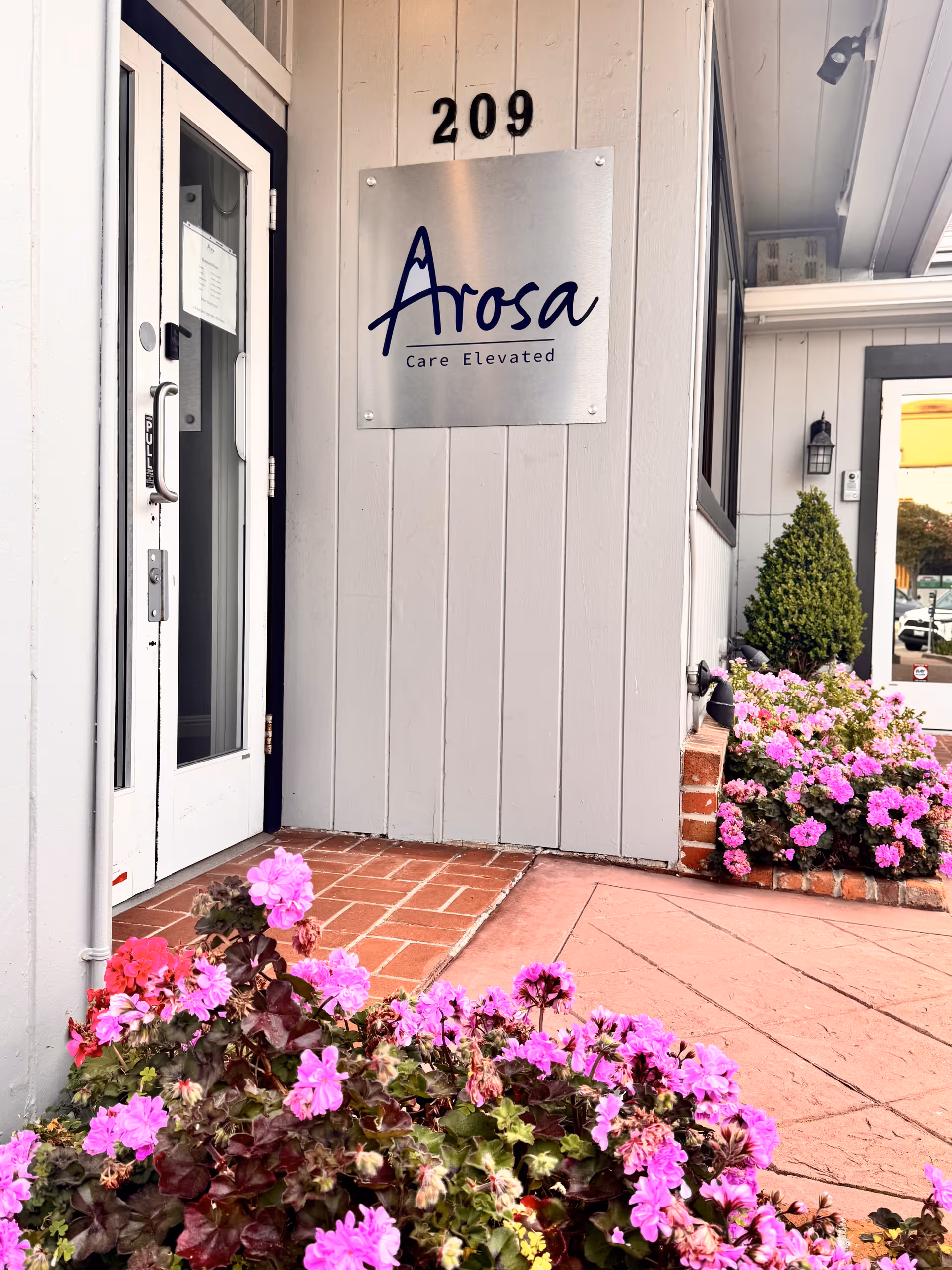 Arosa senior living exterior entrance with signage, brick walkway, and pink flowers.