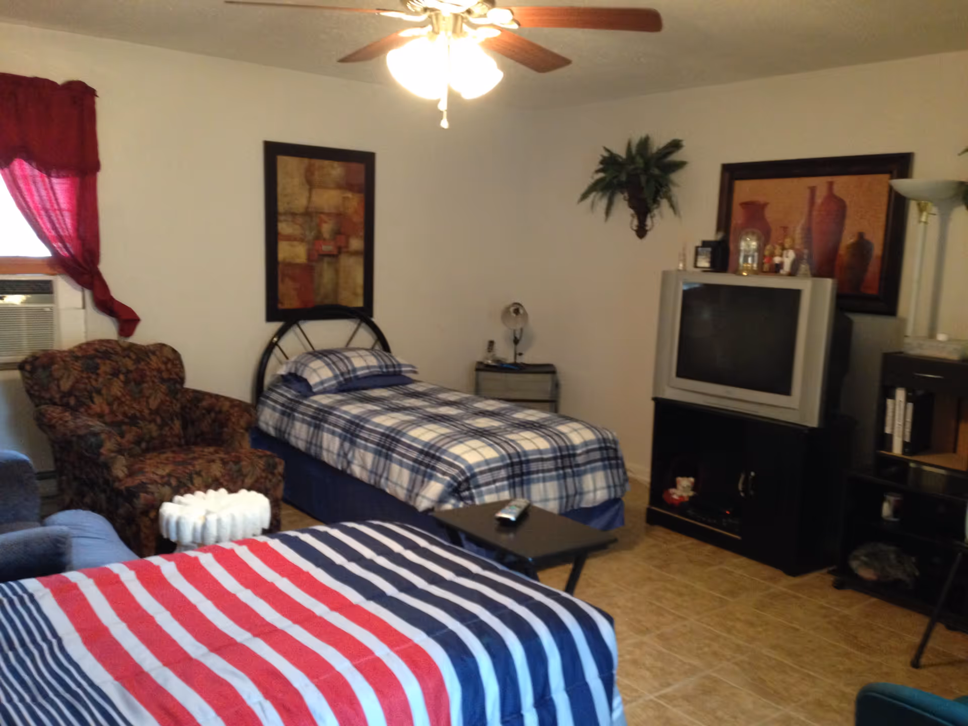 Shared bedroom with two single beds, an armchair, a TV on a stand, and wall decor under a ceiling fan.