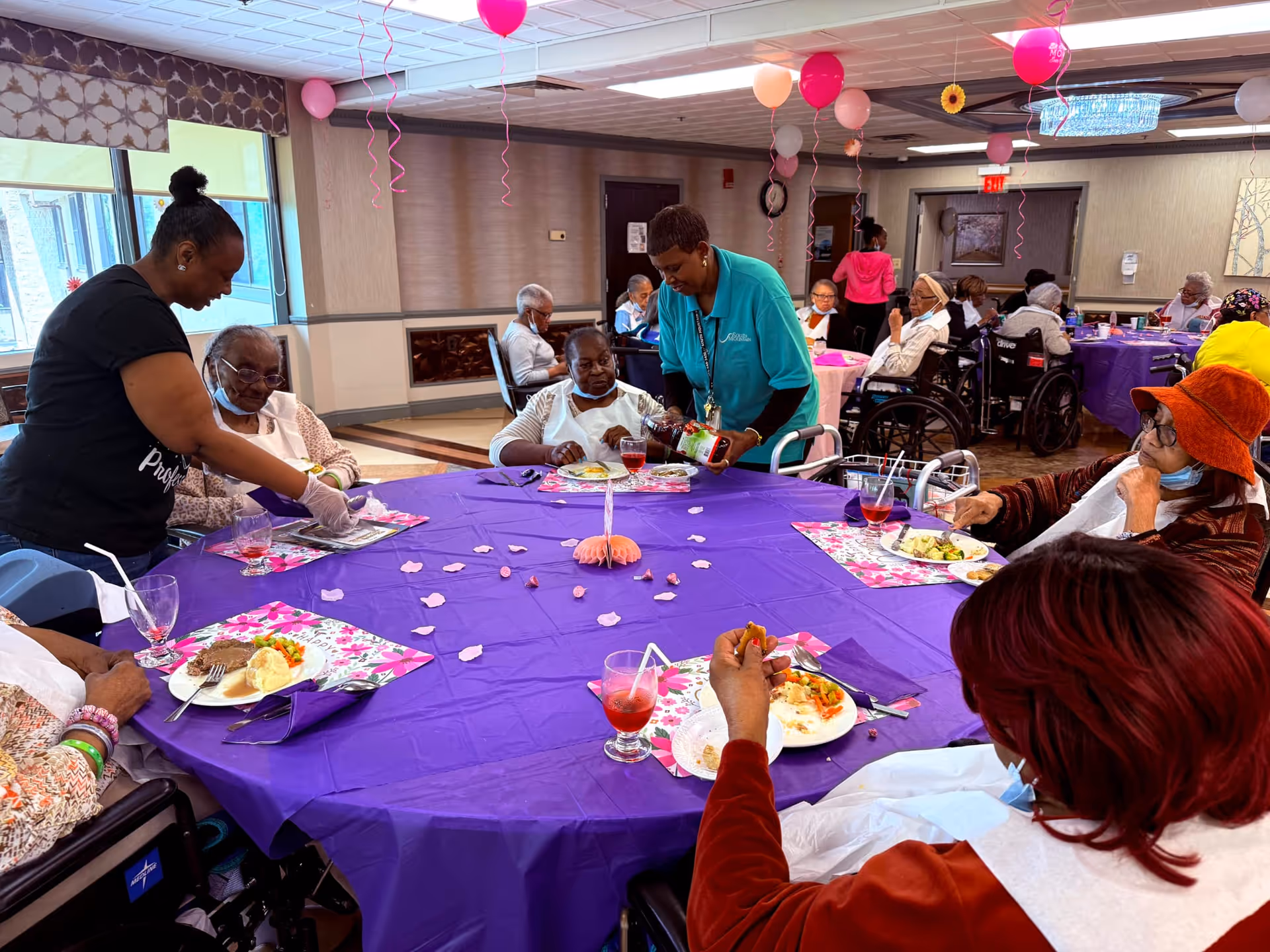 A group of elderly people seated around a large round table covered with a purple tablecloth in a decorated dining room. Two caregivers are assisting them, one serving food and the other pouring a drink. The room is decorated with pink and red balloons hanging from the ceiling, and the elderly individuals are enjoying a meal together.
