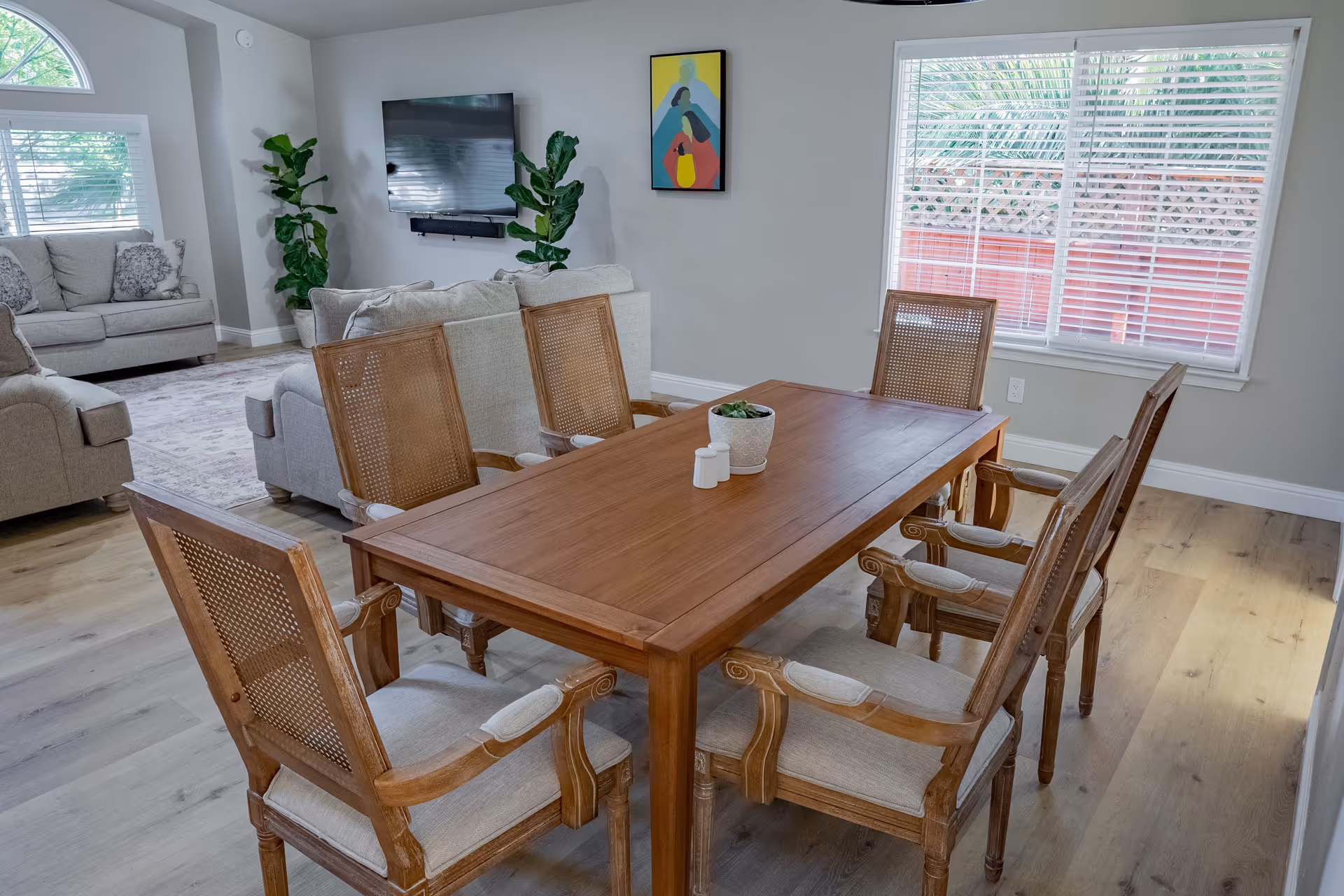 A bright and cozy living and dining area in a care home. The foreground features a wooden dining table with six cushioned wooden chairs. On the table, there is a small potted plant and salt and pepper shakers. In the background, there are two beige sofas with decorative pillows, a wall-mounted flat-screen TV, two tall green plants, and a colorful abstract painting on the wall. Large windows with white blinds allow natural light to fill the room, highlighting the light wood flooring.