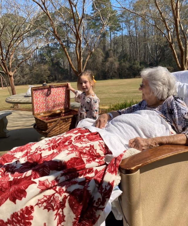 Elderly woman relaxing in an outdoor chair with a young girl and a picnic basket nearby.