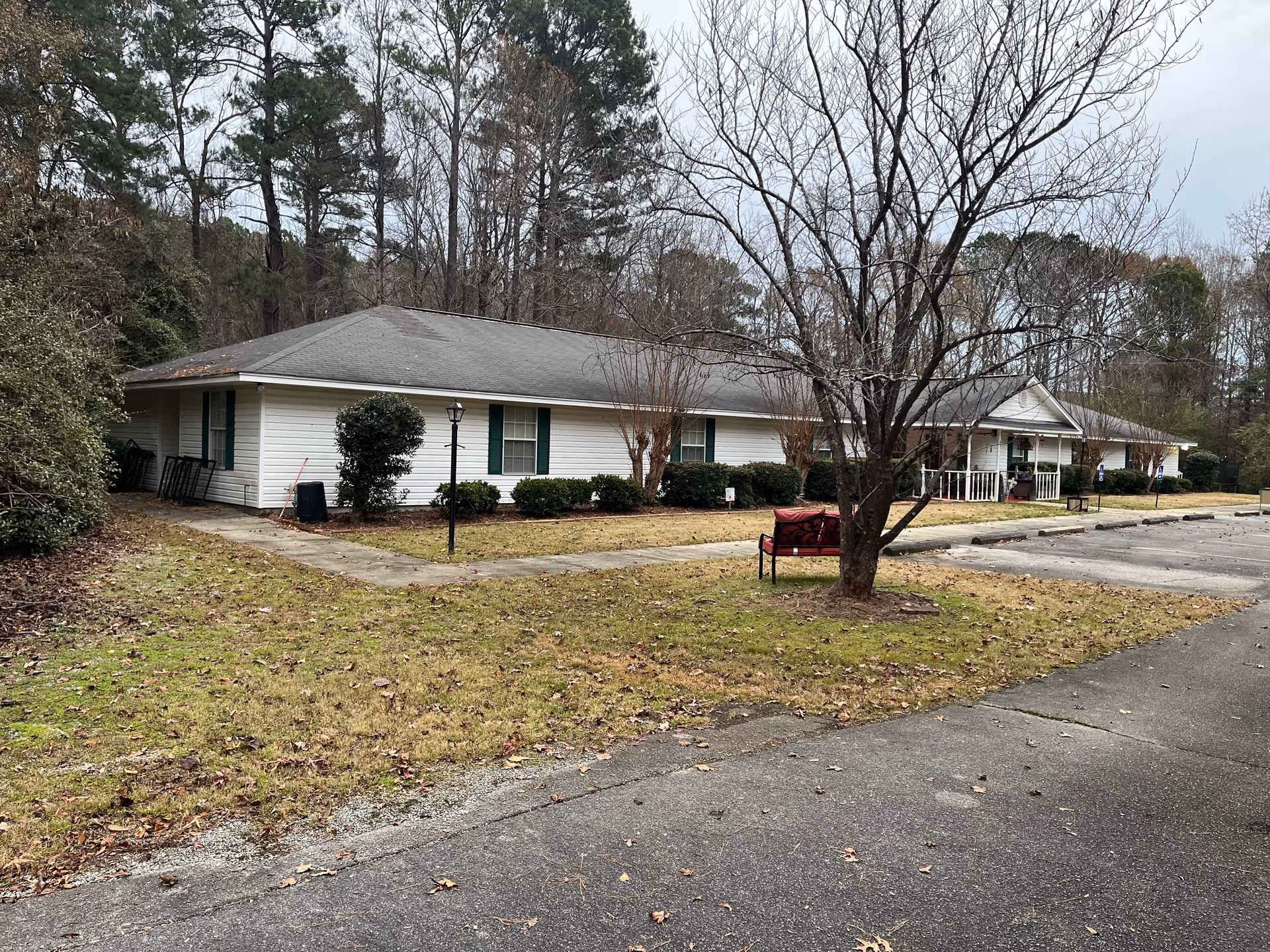 Exterior view of a single-story white building with green shutters, surrounded by trees and bushes. There is a paved parking area to the right and a grassy area with a leafless tree and a red bench in the foreground. The sky is overcast.