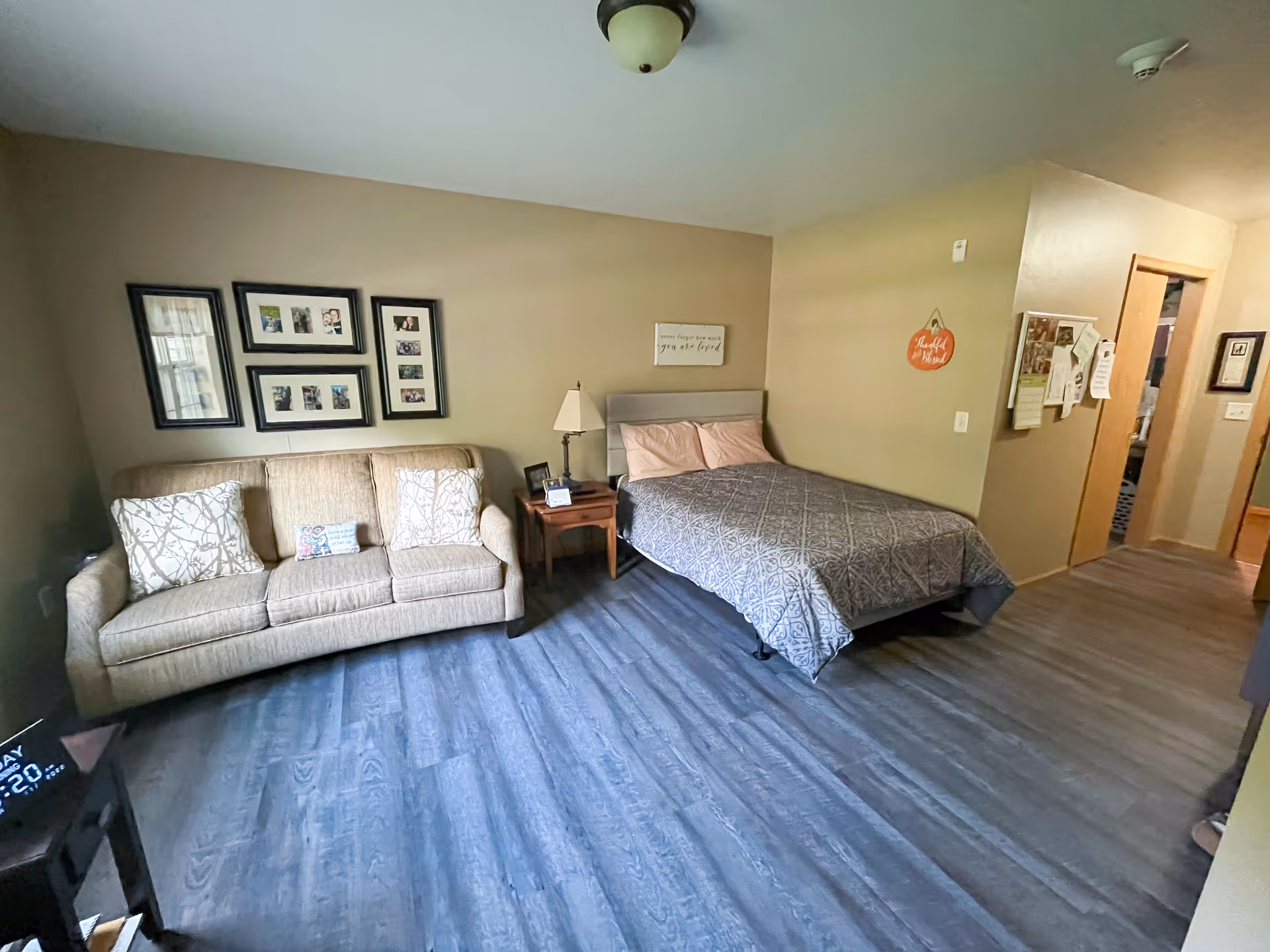 A cozy bedroom with a beige sofa on the left side adorned with patterned pillows, a bed with a gray patterned bedspread and two pillows, a wooden nightstand with a lamp and picture frame, and several framed photos on the wall above the sofa. The room has wood flooring and beige walls with a small decorative sign hanging near the doorway.