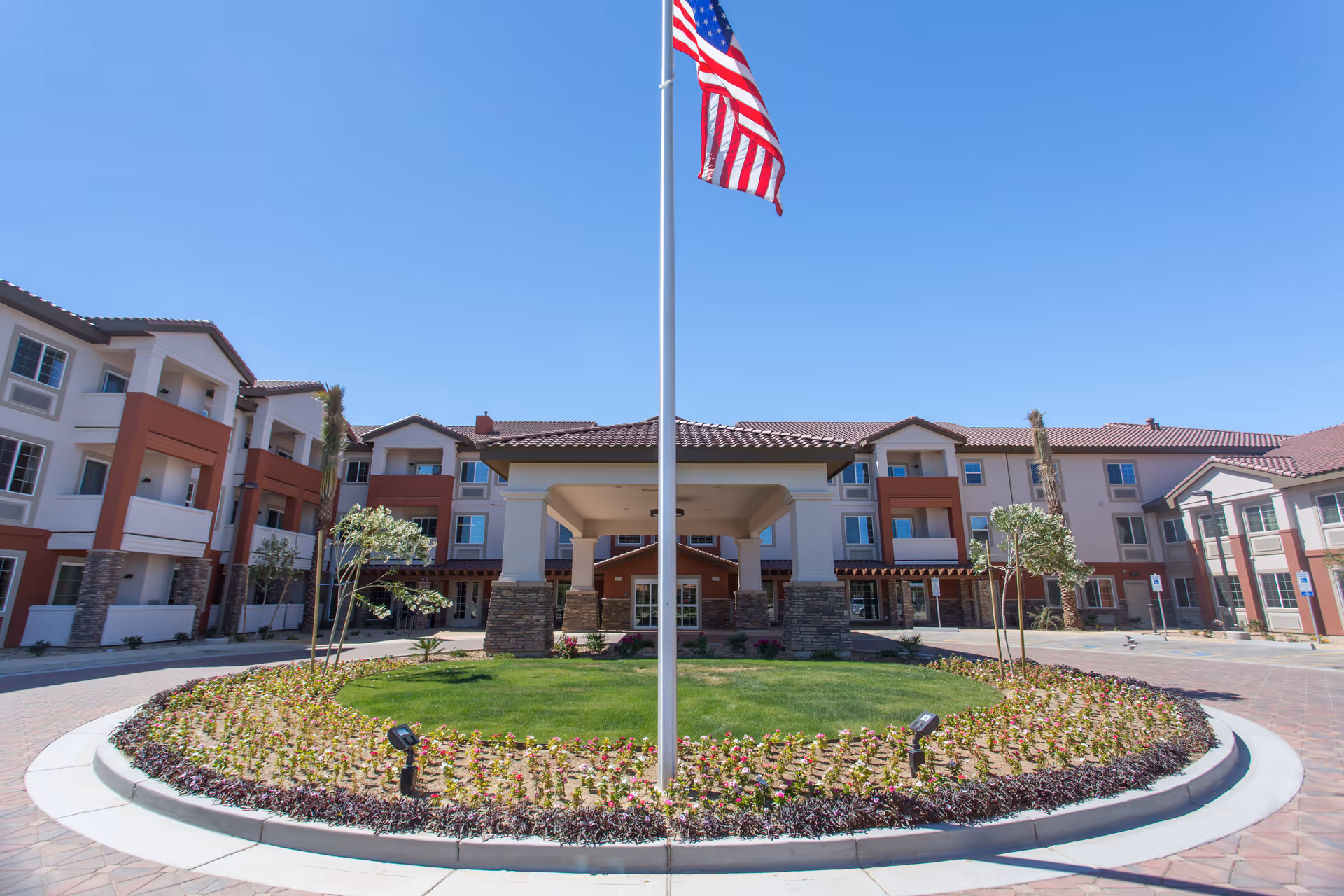 Front view of a senior living campus with a circular garden, flagpole, and surrounding buildings.