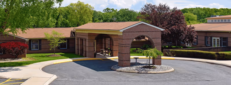 Exterior view of Forest Hill Heights Senior Living facility showing a brick building with a covered entrance and a circular driveway surrounded by landscaped greenery and trees.