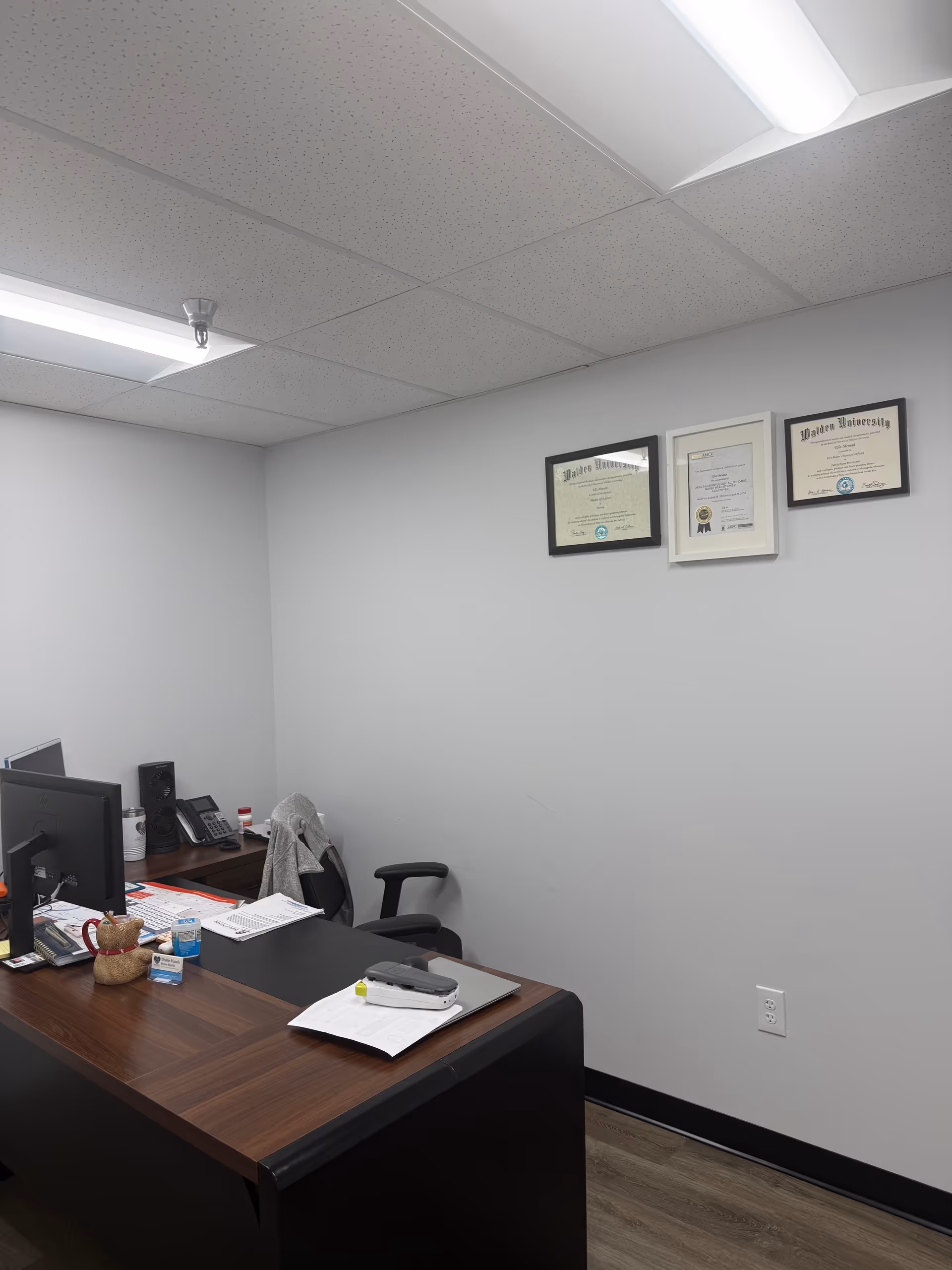 Office desk with computer, certificates on wall, and scattered papers in a small room.