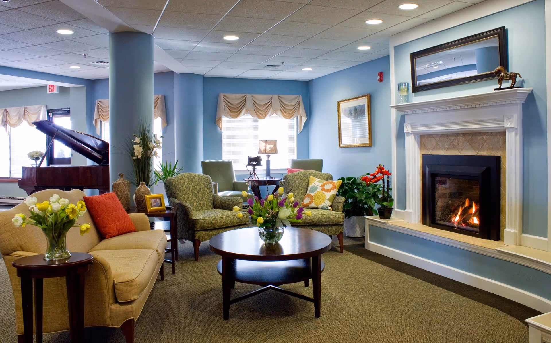 A cozy living room area with a beige sofa adorned with a red pillow, two patterned armchairs, a round wooden coffee table with a vase of flowers, and a lit fireplace with a white mantel. The walls are painted light blue, and there are windows with beige valances letting in natural light. A grand piano is visible in the background near the windows.