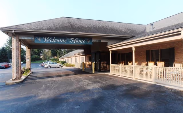 Covered driveway and front entrance of a brick retirement community building with a 'Welcome Home' banner, rocking chairs on the porch, and parked cars.